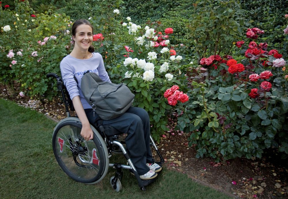 A woman in a purple shirt with a bag in her lap sits in a wheelchair next to a row of flowers, she smiles up at the camera. She is at Hayden Lake, one of our locations.