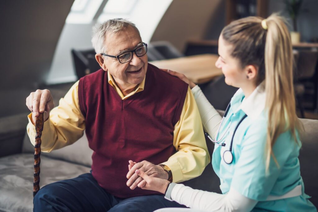 An older man with a can sitting next to a female caretaker who is holding his hand and chatting with him at Basset Creek, one of our locations